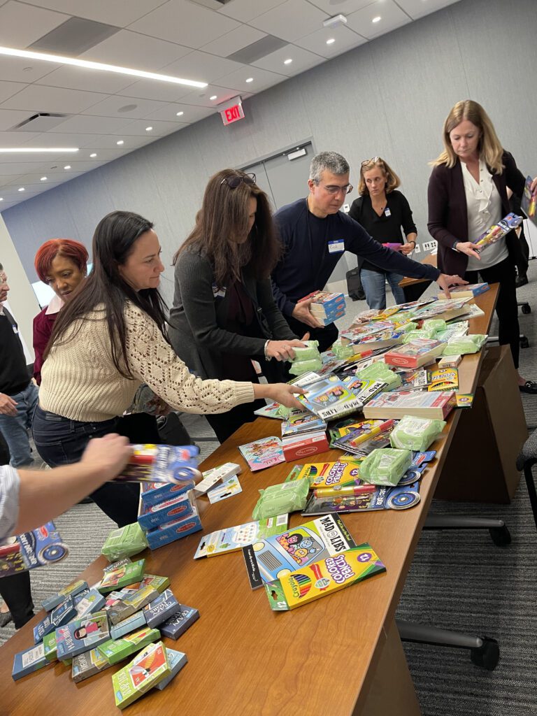 A group of individuals gather around a table filled with various school supplies, including pencils, erasers, and notebooks. They are sorting and organizing the supplies, as part of a team building CSR activity aimed at creating care packages for cancer patients.