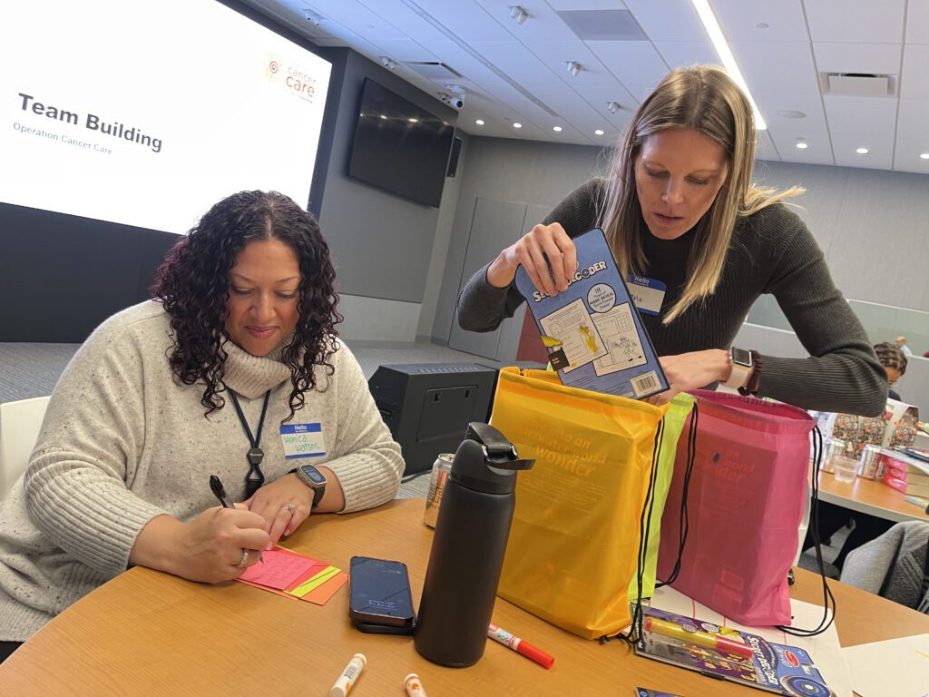 Two women participate in a team building activity as part of the 'Operation Cancer Care' event. One woman is writing a heartfelt note on colorful post-its, while the other is carefully packing a drawstring bag with puzzle books and supplies. Various materials, such as markers, a phone, and a water bottle, are spread across the table. A large screen in the background displays 'Team Building' and 'Operation Cancer Care,' adding context to the charitable activity.