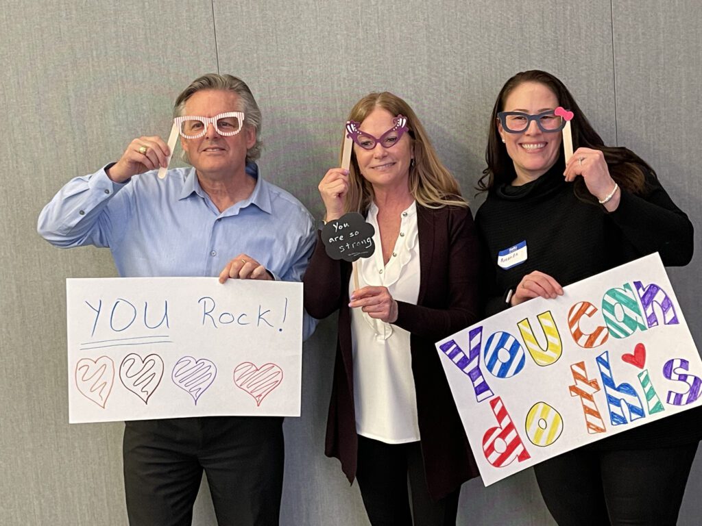 Three individuals holding up handmade motivational signs and wearing playful glasses during a team building event. The signs say 'YOU Rock!' with colorful hearts and 'You can do this,' encouraging messages meant to uplift and inspire.