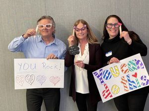 Three individuals holding up handmade motivational signs and wearing playful glasses during a team building event. The signs say 'YOU Rock!' with colorful hearts and 'You can do this,' encouraging messages meant to uplift and inspire.