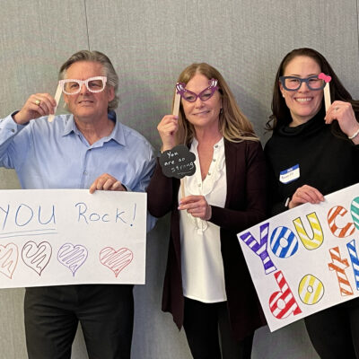 Three individuals holding up handmade motivational signs and wearing playful glasses during a team building event. The signs say 'YOU Rock!' with colorful hearts and 'You can do this,' encouraging messages meant to uplift and inspire.