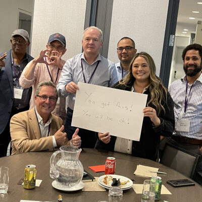 A group of seven people standing and seated around a table during a team building event. One woman holds a white sign that reads, 'You got this! You will be ok!' Others are smiling and giving thumbs up, showing supportive gestures.