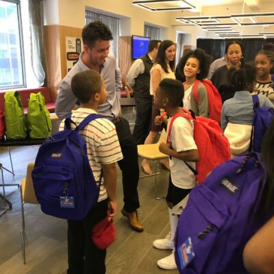 A team building participant interacts with children during a Tools for Schools CSR event, as they receive brightly colored backpacks filled with school supplies.