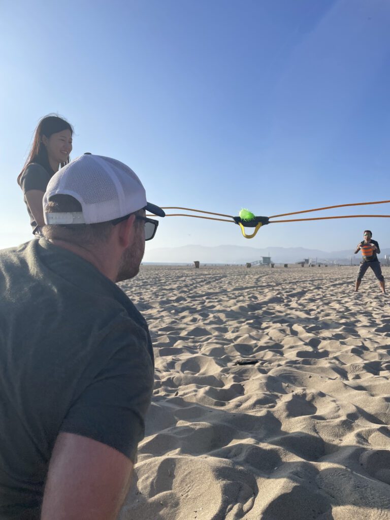 Team members use a slingshot during a Corporate Survivor beach activity, focusing on strategy and team coordination.