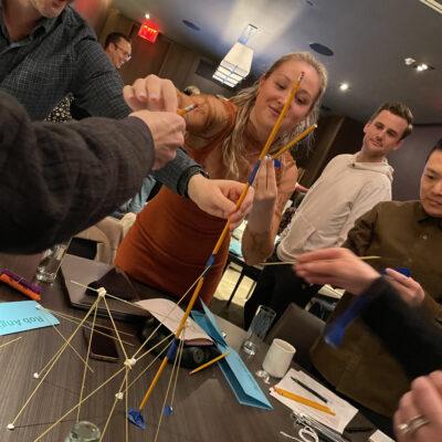 Group of colleagues participating in a team building activity, collaborating to construct a tall structure using spaghetti and marshmallows; and through that challenge win school supplies to donate to students.