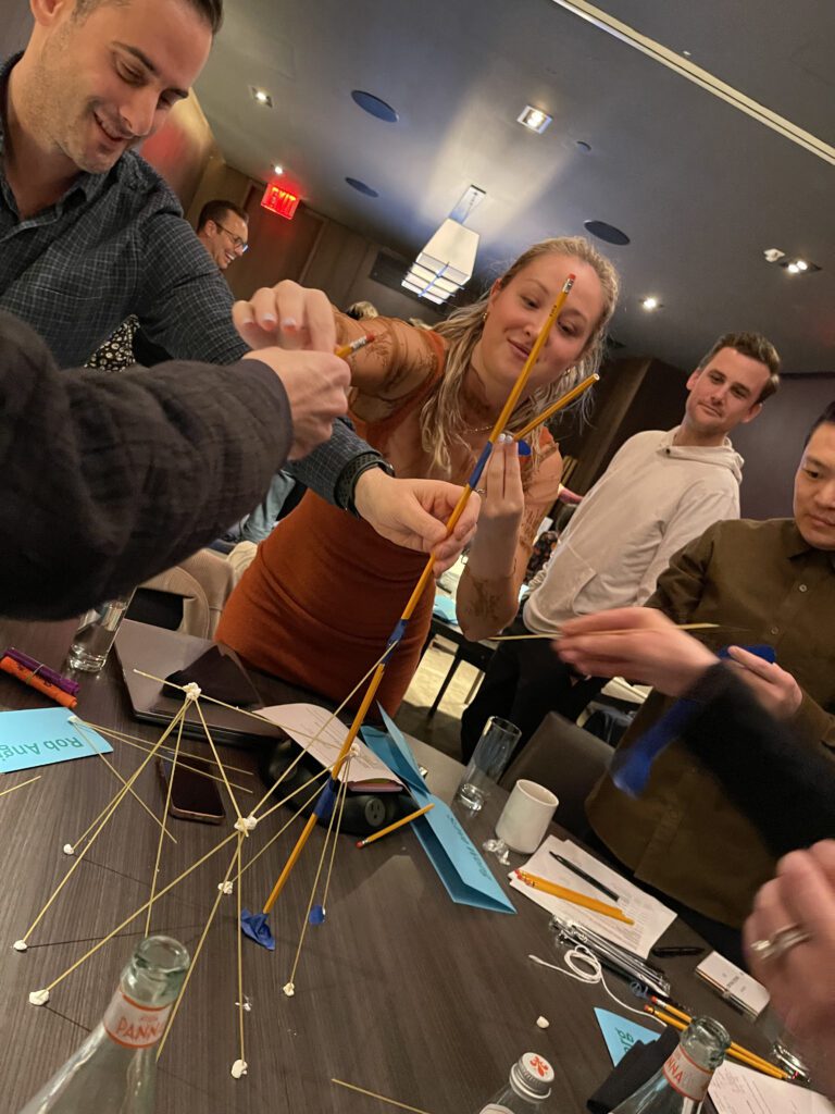 Group of colleagues participating in a team building activity, collaborating to construct a tall structure using spaghetti and marshmallows; and through that challenge win school supplies to donate to students.