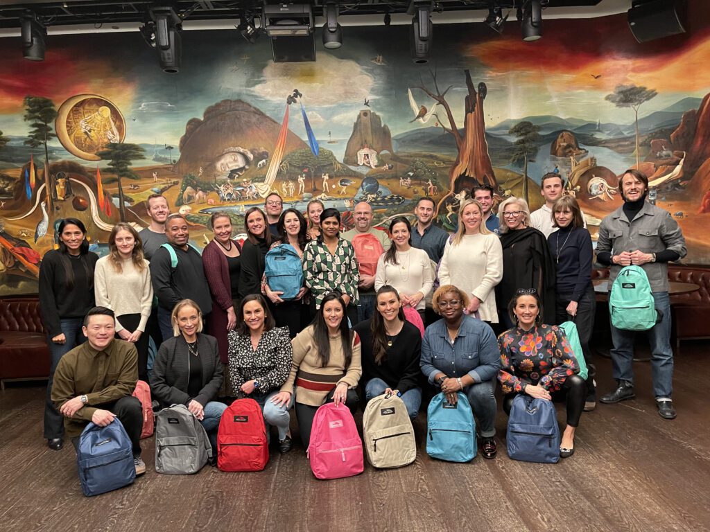 Large group of colleagues standing together, proudly displaying colorful backpacks they have packed during a team building CSR event.