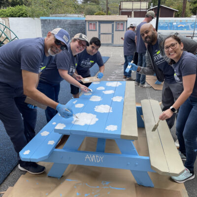 A group of five volunteers working together to paint an outdoor picnic table at a charitable team building event. The table is painted in bright blue with white clouds. All participants, wearing matching grey shirts and blue gloves, are smiling as they contribute to the project.