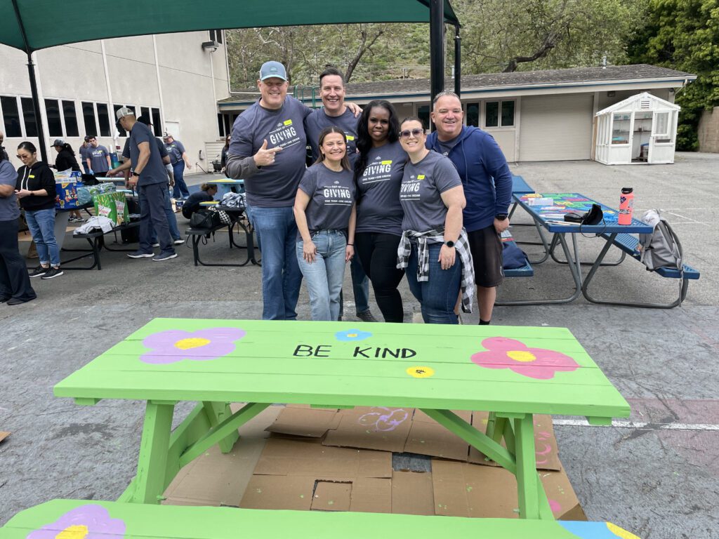 A group of six people standing together, smiling in front of a brightly painted picnic table that reads 