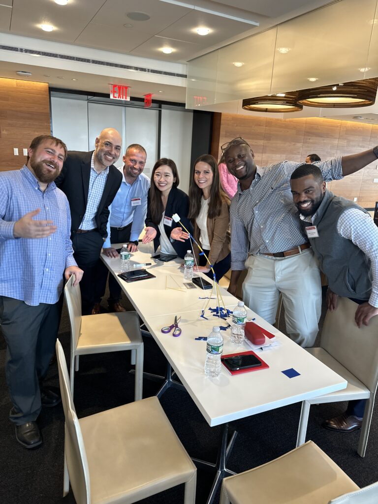 A cheerful group of coworkers posing together around a table after completing a team building activity with marshmallows and spaghetti towers.