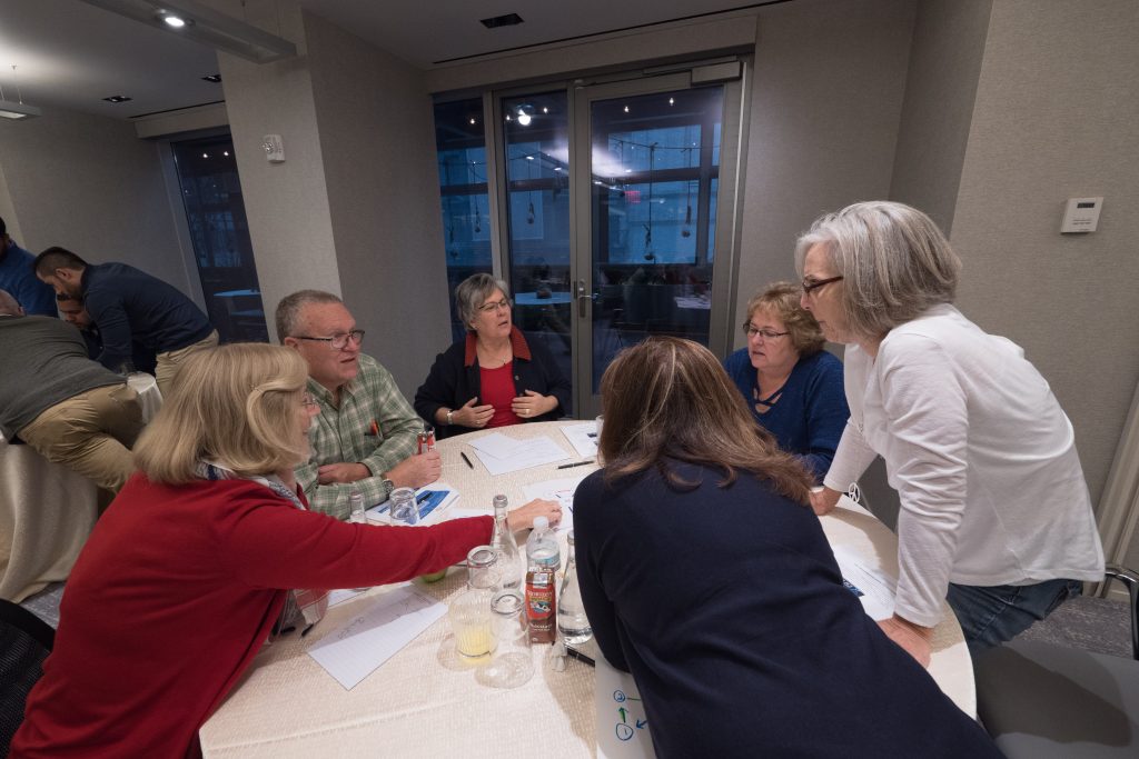 A group of participants collaborating at a table during a Team DNA workshop, discussing and working on materials with a facilitator nearby.