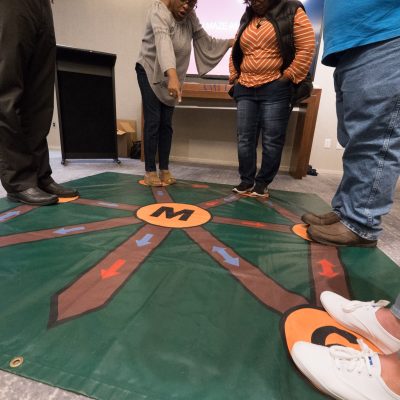 Participants engaging in an interactive Team and Leadership DNA activity, using a large, colorful floor mat designed to explore key team dynamics and enhance communication.