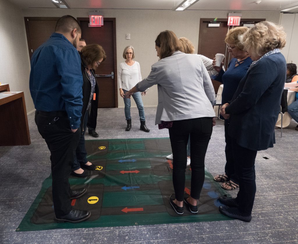 A group participating in a Team DNA activity, standing on a large interactive mat with color-coded arrows, while a facilitator provides instructions.