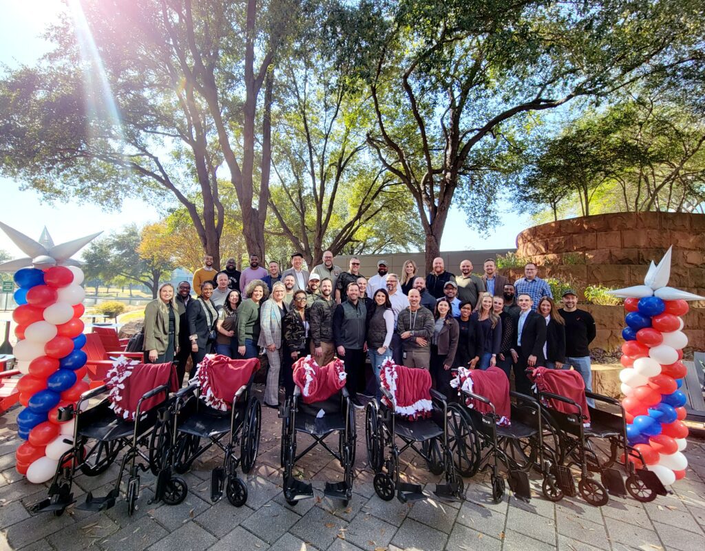 A large group of participants stands outdoors behind a row of assembled wheelchairs decorated with patriotic red, white, and blue ribbons during the Wheelchairs In Motion team building event. The group is celebrating their successful charitable effort.