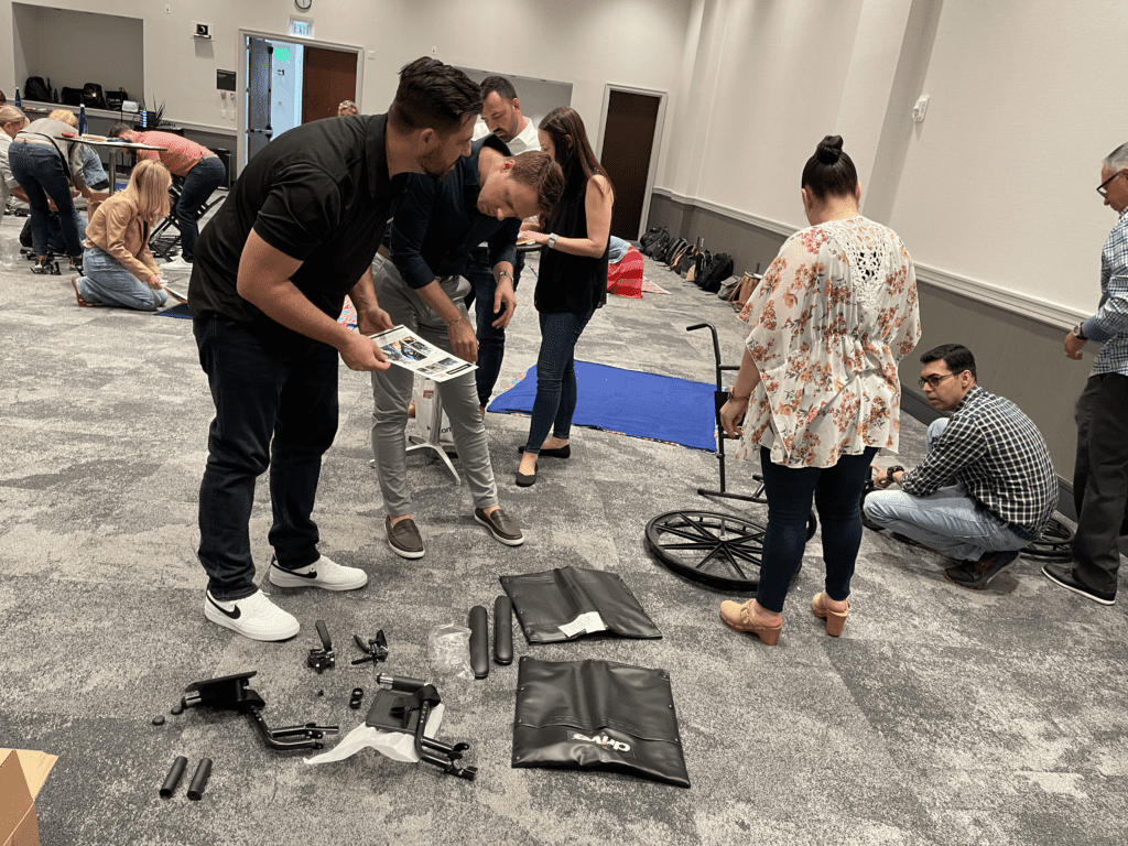 Participants gather around wheelchair parts, working together to assemble them during the Wheelchairs In Motion team building event. The room is filled with groups collaborating on similar tasks as part of the charitable activity.