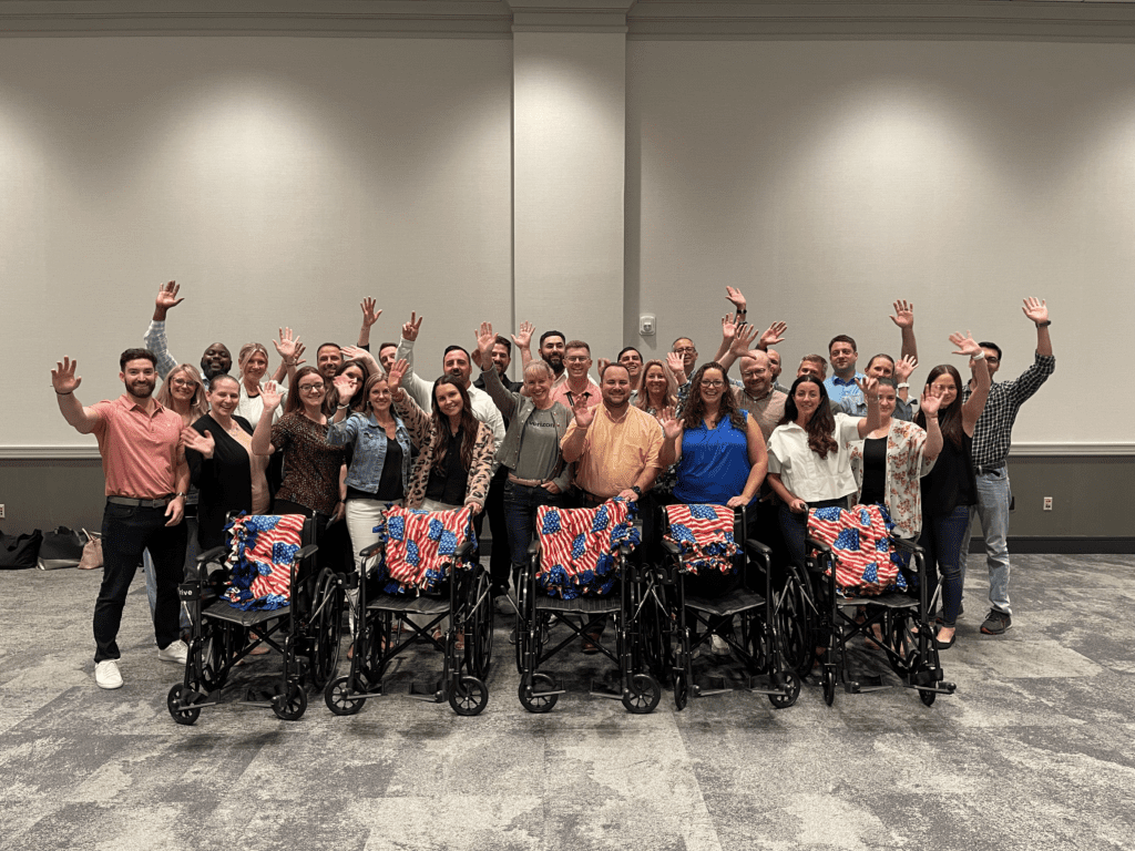 A large group of participants stands behind a row of assembled wheelchairs decorated with American flag blankets, celebrating their achievement during the Wheelchairs In Motion team building event. The group is smiling and raising their hands in celebration.