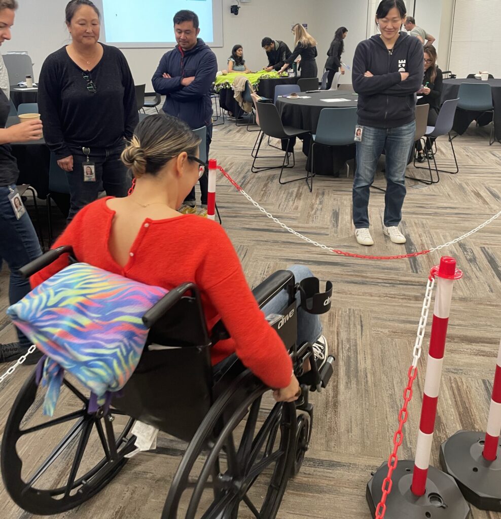 A participant navigates an obstacle course in a wheelchair during the Wheelchairs In Motion team building event. Other participants watch and support as she tests the newly assembled wheelchair.