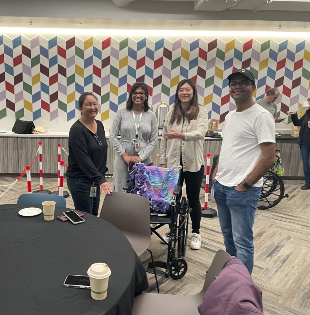 A group of participants gathers around a table in a colorful indoor space during the Wheelchairs In Motion team building event. A decorated wheelchair is nearby, and the team is smiling, enjoying the charitable activity.