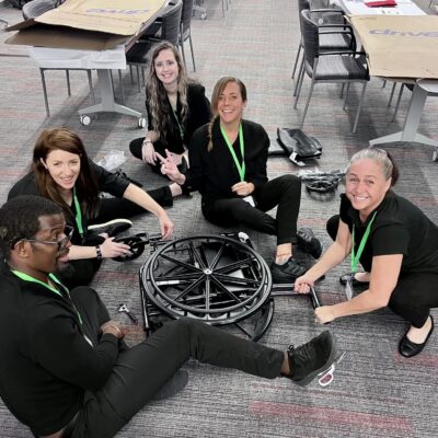 A group of smiling participants sits on the floor working together to assemble a wheelchair during the Wheelchairs In Motion team building event. The activity is part of a charitable initiative to donate wheelchairs to those in need.