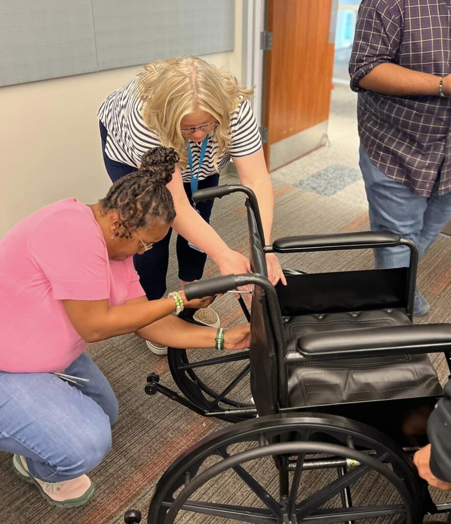 Two participants work together to assemble a wheelchair during the Wheelchairs In Motion team building event. They focus on adjusting and securing parts of the wheelchair frame as part of the charitable activity.