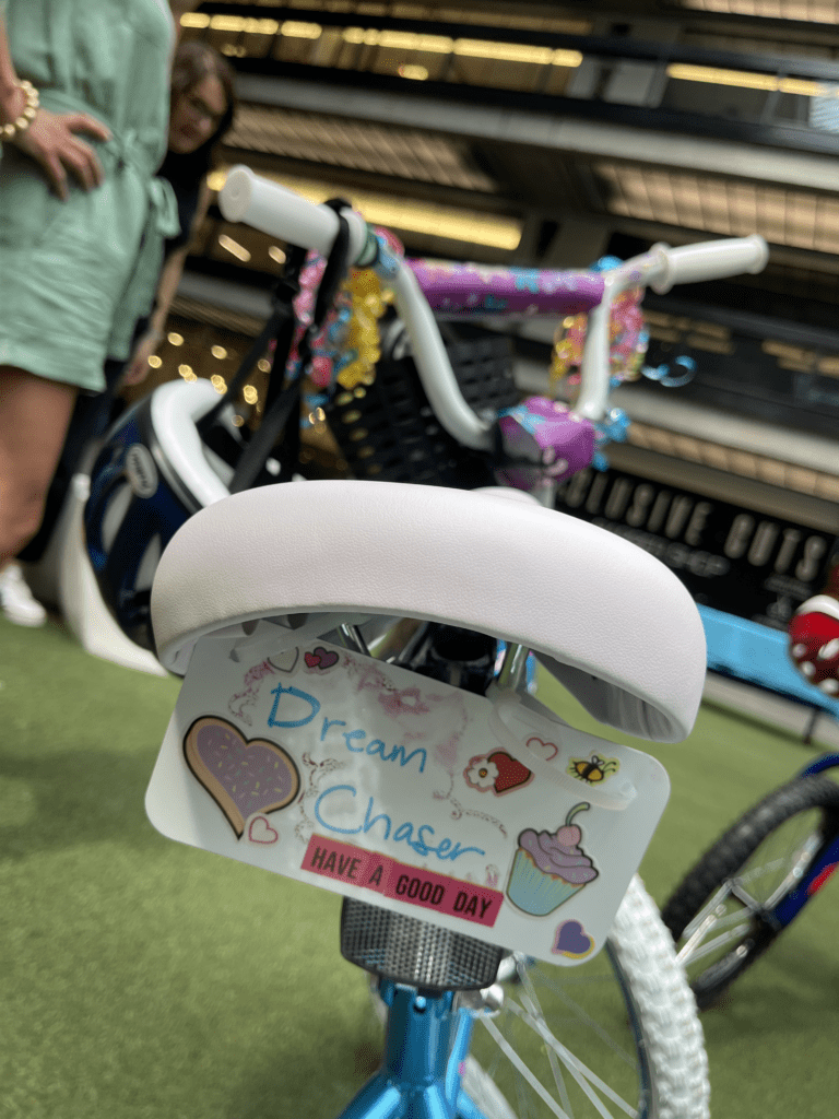 Close-up of a decorated bicycle from a Charity Bike Build event. The bike seat features a personalized tag reading 'Dream Chaser' with colorful stickers, including hearts, a cupcake, and motivational text 'Have a Good Day,' showing thoughtful details in the bike assembly process.