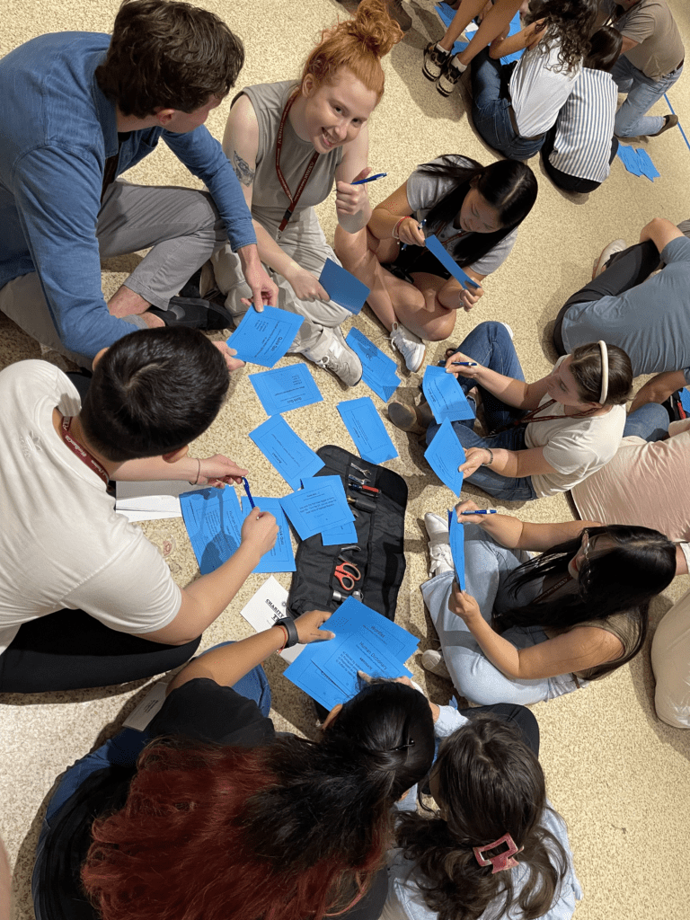 A group of people sitting in a circle on the floor during a team building activity, each holding blue cards with instructions. They are engaging in a collaborative task, exchanging ideas and writing down notes.