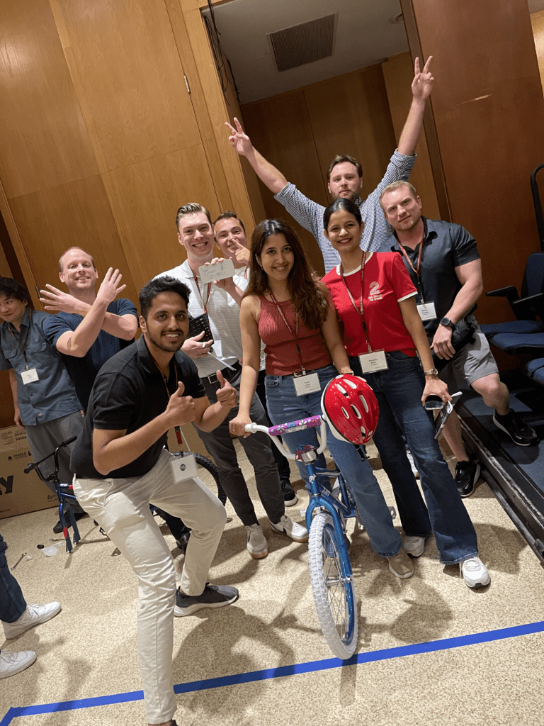 A group of smiling participants posing proudly with a completed bicycle during a Charity Bike Build event. Some team members are giving thumbs up and raising their hands in excitement, celebrating their achievement after assembling the bike.