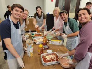 A group of six participants in the 'Cooking For A Cause' team building event smiling as they prepare meals to donate. They are gathered around a wooden table, each wearing plastic aprons and gloves while assembling dishes in aluminum trays. The team is working collaboratively, spreading sauce and layering ingredients, likely for a lasagna or similar dish. This activity combines teamwork and community service by providing meals for local food banks or soup kitchens.