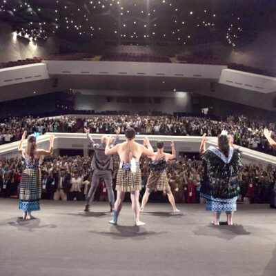 Maori performers in traditional attire lead a large audience in an energetic Haka team building session on a grand stage, inspiring unity and passion.