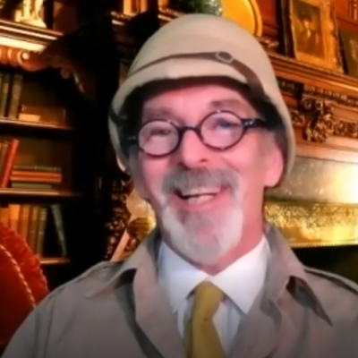 A man with glasses and a safari-style pith helmet smiles warmly in front of a cozy, vintage room filled with bookshelves. He is dressed in character, wearing a light-colored jacket and a yellow tie. The man is in character, part of a role-playing murder mystery team building event.