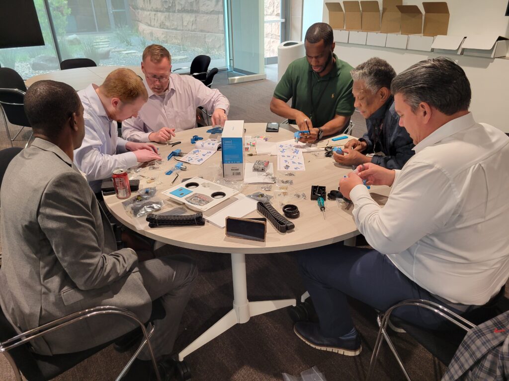 A group of six individuals is seated around a circular table, deeply engaged in assembling small robotic kits. Various parts, tools, and instruction manuals are spread out across the table. The participants are focused on the task, collaborating as they build robots for a team building activity.