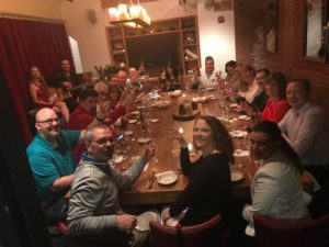 A large group of people gathered around a long wooden table in a warmly lit dining room for a Murder Mystery Dinner event. Each person is holding a glass of wine, smiling, and raising their glass in a celebratory toast. The table is set with elegant dinnerware and a centerpiece featuring a decorative cake. The atmosphere is festive, with participants ready to enjoy both the meal and the interactive murder mystery experience.