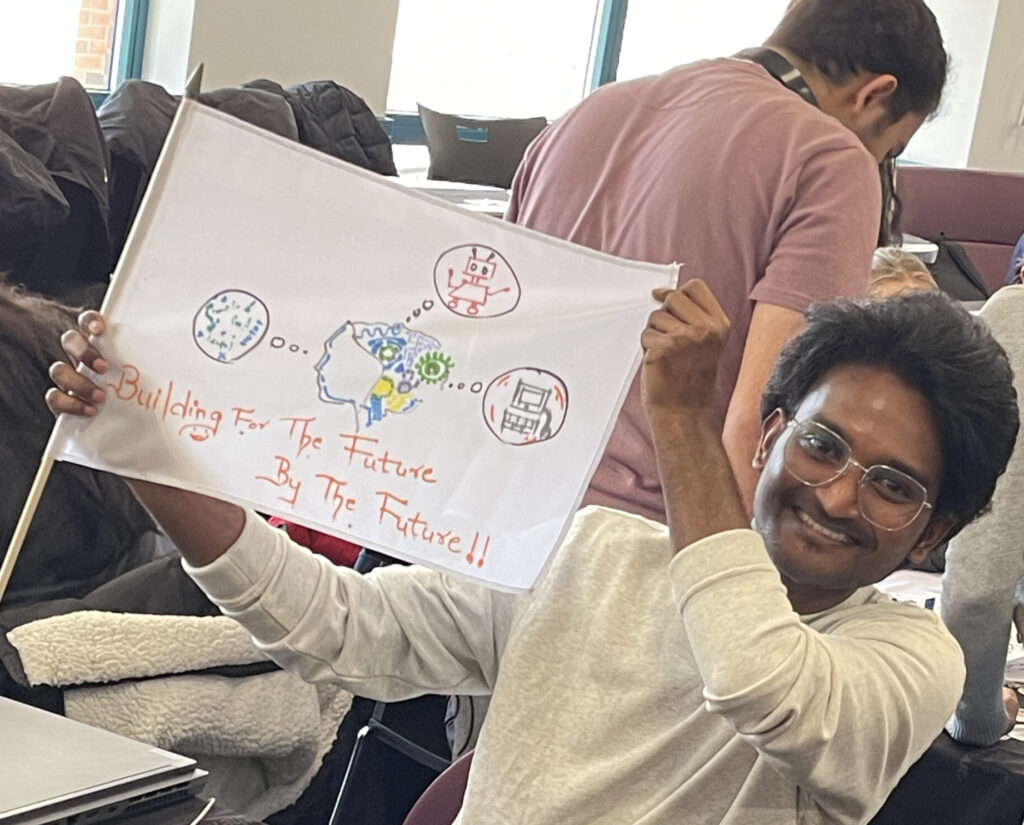 A young man is proudly holding up a flag he created, which features hand-drawn images of technology-related elements like a robot and text that reads, 