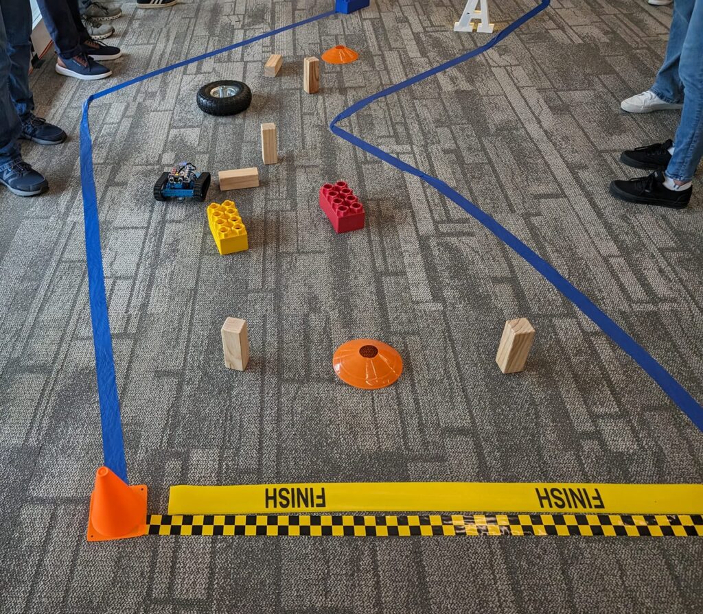 A small robot is navigating through an obstacle course marked by blue tape on the floor. Various objects, such as large toy bricks, wooden blocks, and traffic cones, create the course's challenges. The 