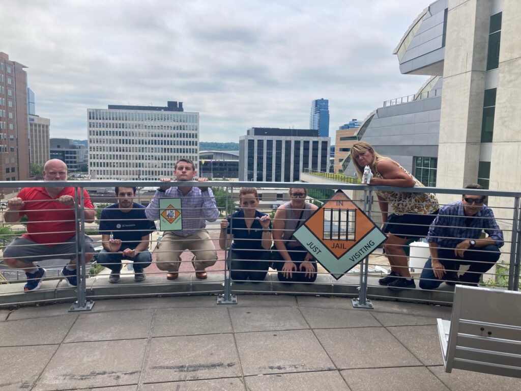 Team members posing behind a railing holding Monopoly-style 'In Jail' and 'Go to Jail' signs during a Team-opoly scavenger hunt team building event, with a cityscape in the background.