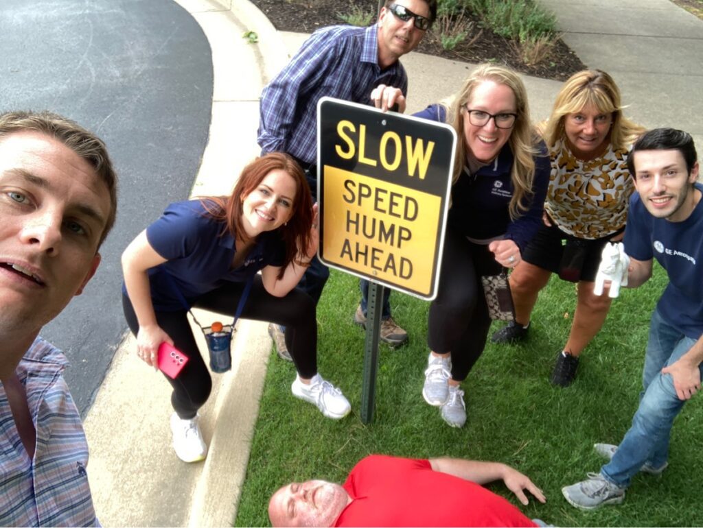 Team gathered around a 'Slow Speed Hump Ahead' sign, with one member humorously lying down next to it during a Team-opoly scavenger hunt team building event.