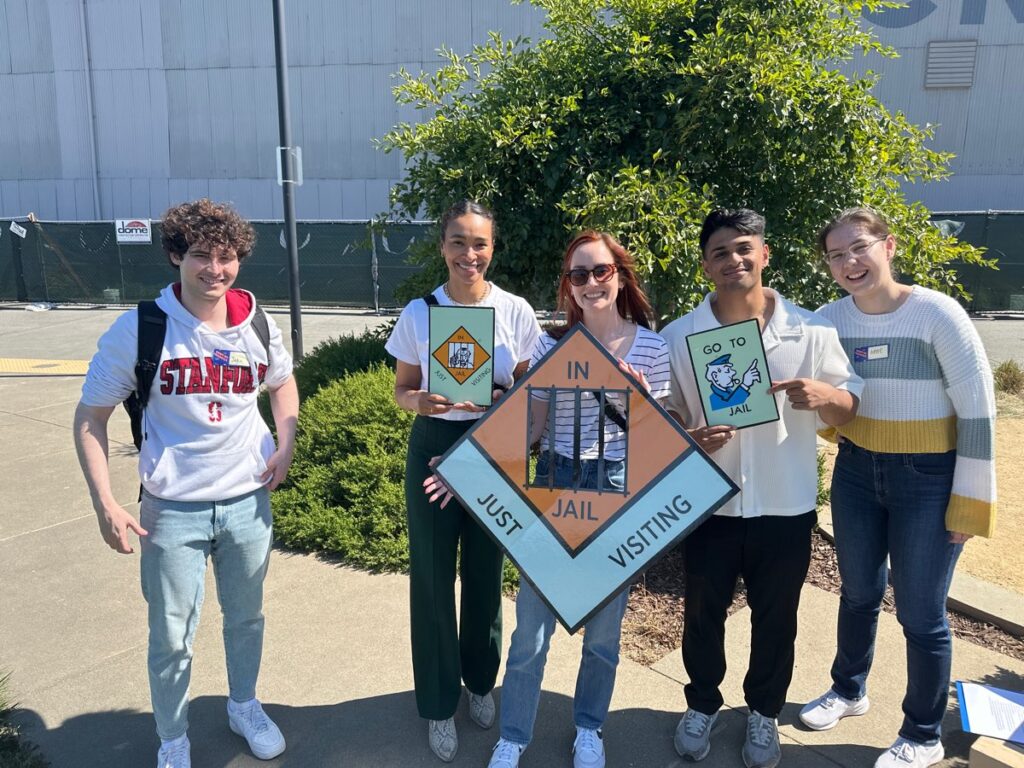 Group of participants posing with Monopoly-style 'In Jail' and 'Go to Jail' signs during a Team-opoly scavenger hunt outdoors, with smiles and playful expressions.