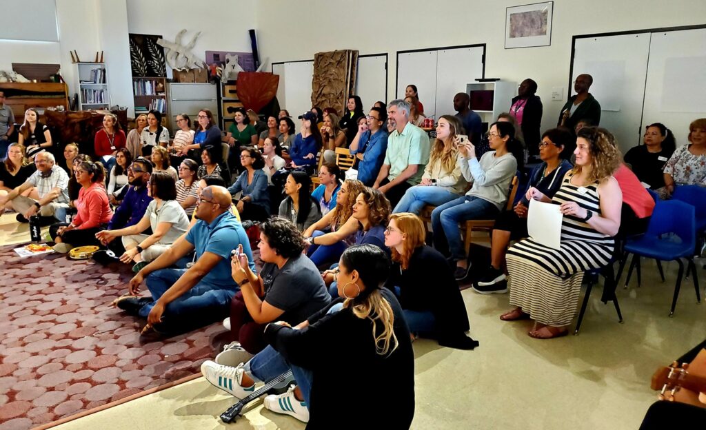 A large group of participants sits and watches a performance during a team building event. They are gathered in a spacious room, engaged and enjoying the presentation as part of the Team Ukulele activity.