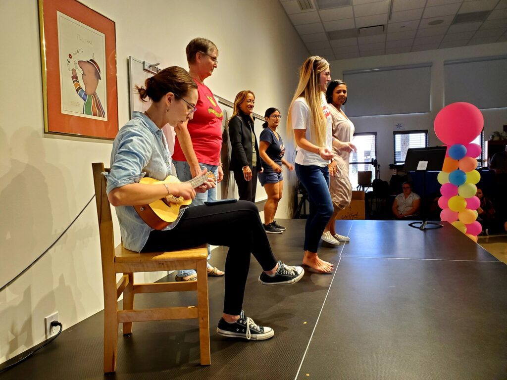 Participants perform on stage during a team building event. One person sits and plays the ukulele while others stand nearby, preparing to sing or dance as part of the Team Ukulele activity.