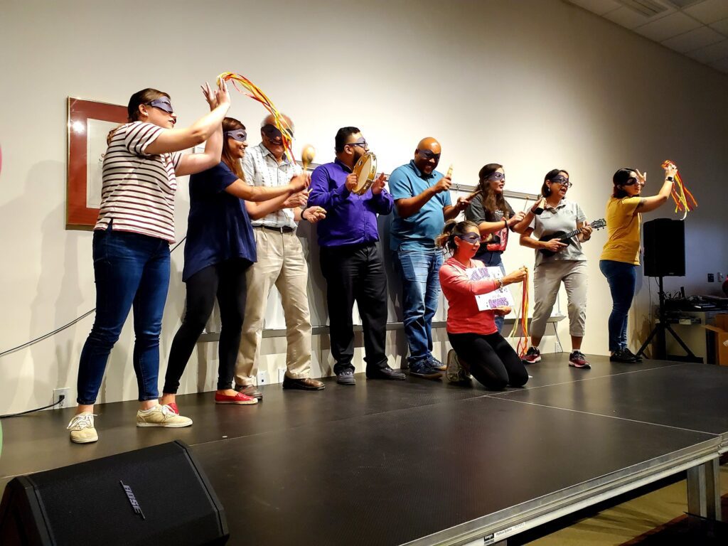 A group of participants stands on stage, wearing blindfolds and holding various props, including ukuleles and ribbons, as part of a team building event. They are engaged in a collaborative performance during the Team Ukulele activity.