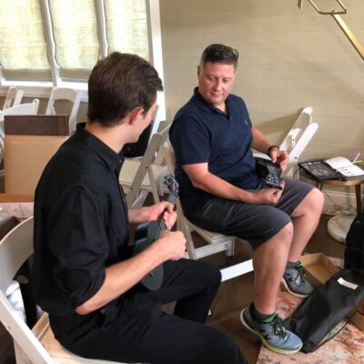 Two participants sitting and holding ukuleles during a team building event. They are engaged in conversation, learning to play their instruments as part of the Team Ukulele music activity.