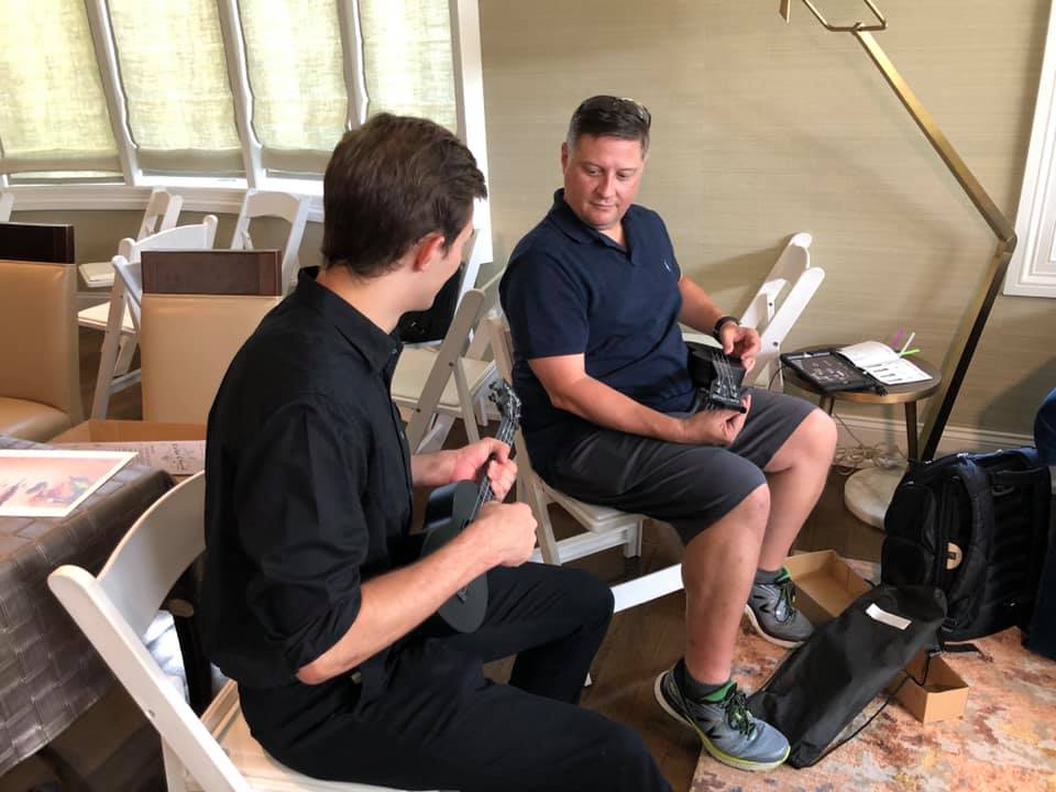 Two participants sitting and holding ukuleles during a team building event. They are engaged in conversation, learning to play their instruments as part of the Team Ukulele music activity.