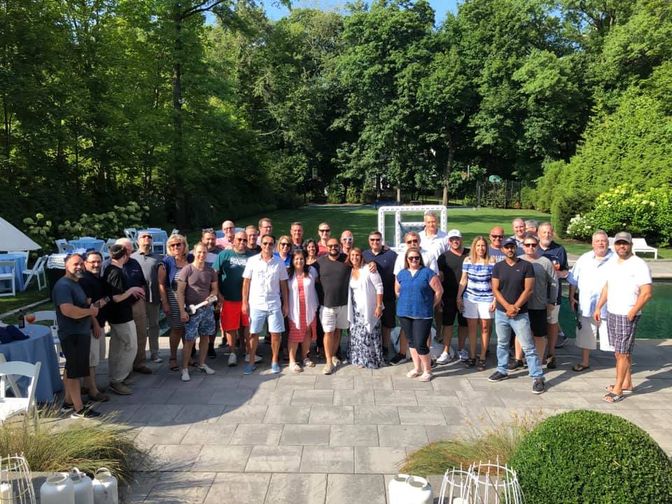 A large group of participants gathers for a group photo outdoors in a scenic garden area during a team building event. The group stands together smiling, enjoying the Team Ukulele activity.