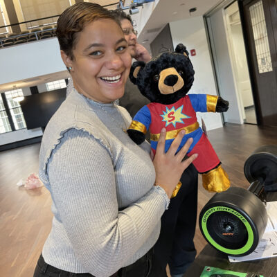 A smiling woman holding a black teddy bear dressed in a superhero outfit, complete with a red suit, gold accents, and a large 'S' emblem on the chest. In the background, a Green Machine bike with neon green wheels is visible.