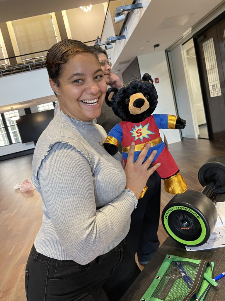 A smiling woman holding a black teddy bear dressed in a superhero outfit, complete with a red suit, gold accents, and a large 'S' emblem on the chest. In the background, a Green Machine bike with neon green wheels is visible.