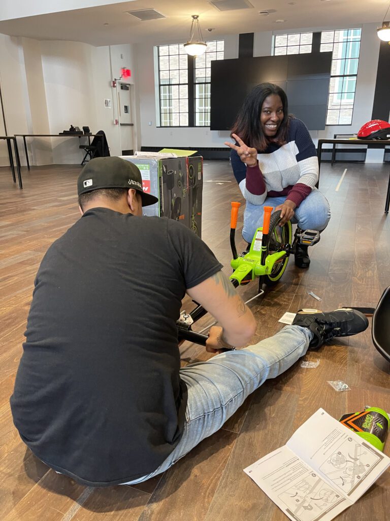 A man sitting on the floor assembling a Green Machine bike as part of a charitable team building event while a woman kneels behind it, flashing a peace sign and smiling.