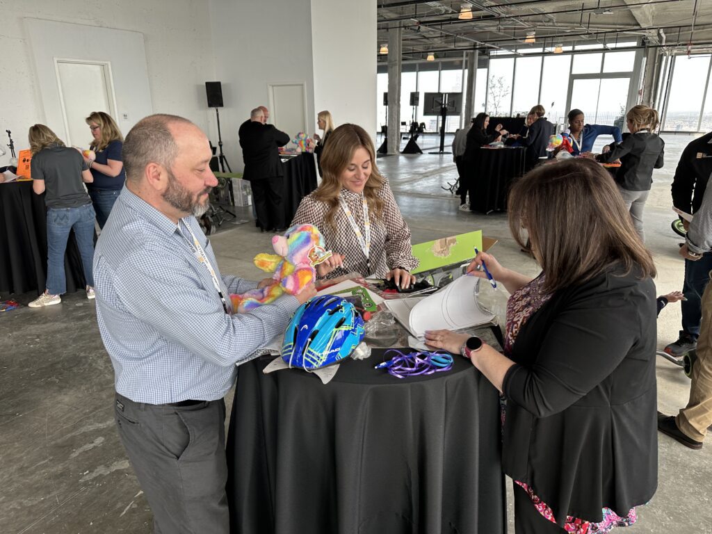 Three people stand around a high-top table, collaborating on assembling items. One person holds a colorful stuffed bear, while another is focused on a helmet and decorations. The third person writes notes on a document. They are participating in a charitable team building activity.