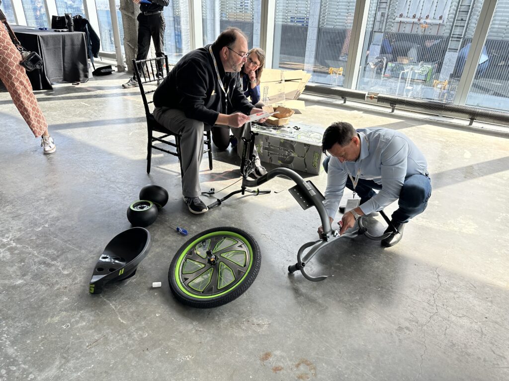 Two people are assembling a Green Machine bike, working together in a well-lit space with large windows. One person is kneeling on the ground, attaching parts of the bike, while the other sits on a chair, reviewing instructions. Bike parts, including the wheel and seat, are spread out on the floor, reflecting a collaborative and charitable team building activity.