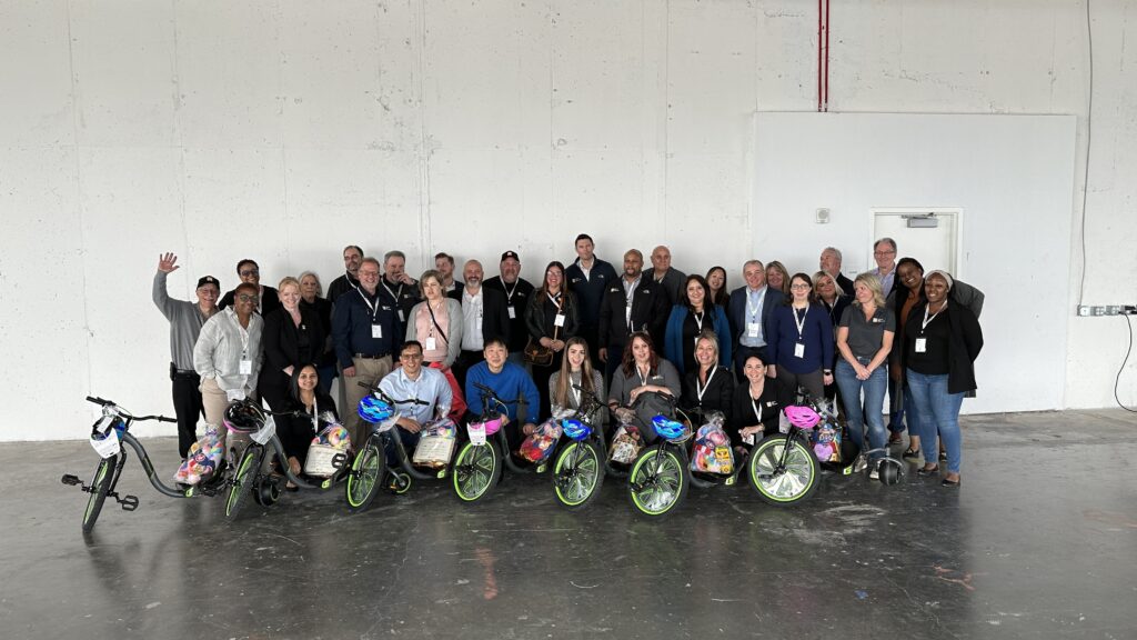 A large group of people poses together indoors, standing behind several Green Machine bikes decorated with colorful baskets and helmets. The group, made up of both seated and standing individuals, is smiling, showing camaraderie and a sense of accomplishment after assembling the bikes during a charitable team building atmosphere.