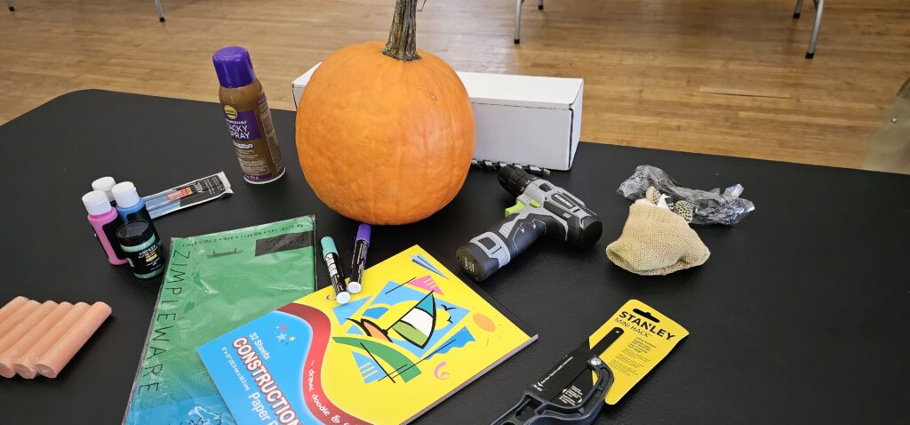 This image shows materials laid out on a table in preparation for assembling a pumpkin racer. A pumpkin sits at the center, surrounded by tools and craft supplies, including a power drill, construction paper, markers, clamps, and paints. These items are essential for designing, decorating, and constructing the pumpkin racer for the upcoming race, creating a fun and creative team building activity.